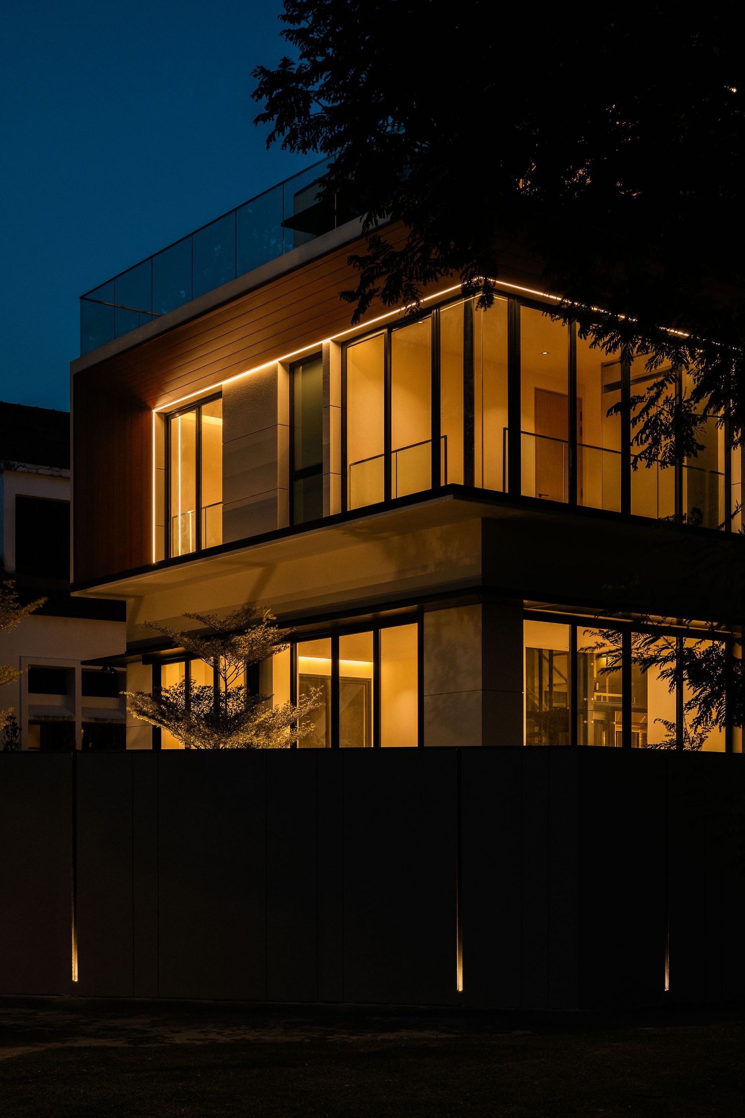 Modern two-story house at dusk with warm glowing windows, wood cladding, glass balcony railing and a tall fence lit by low ground lights.
