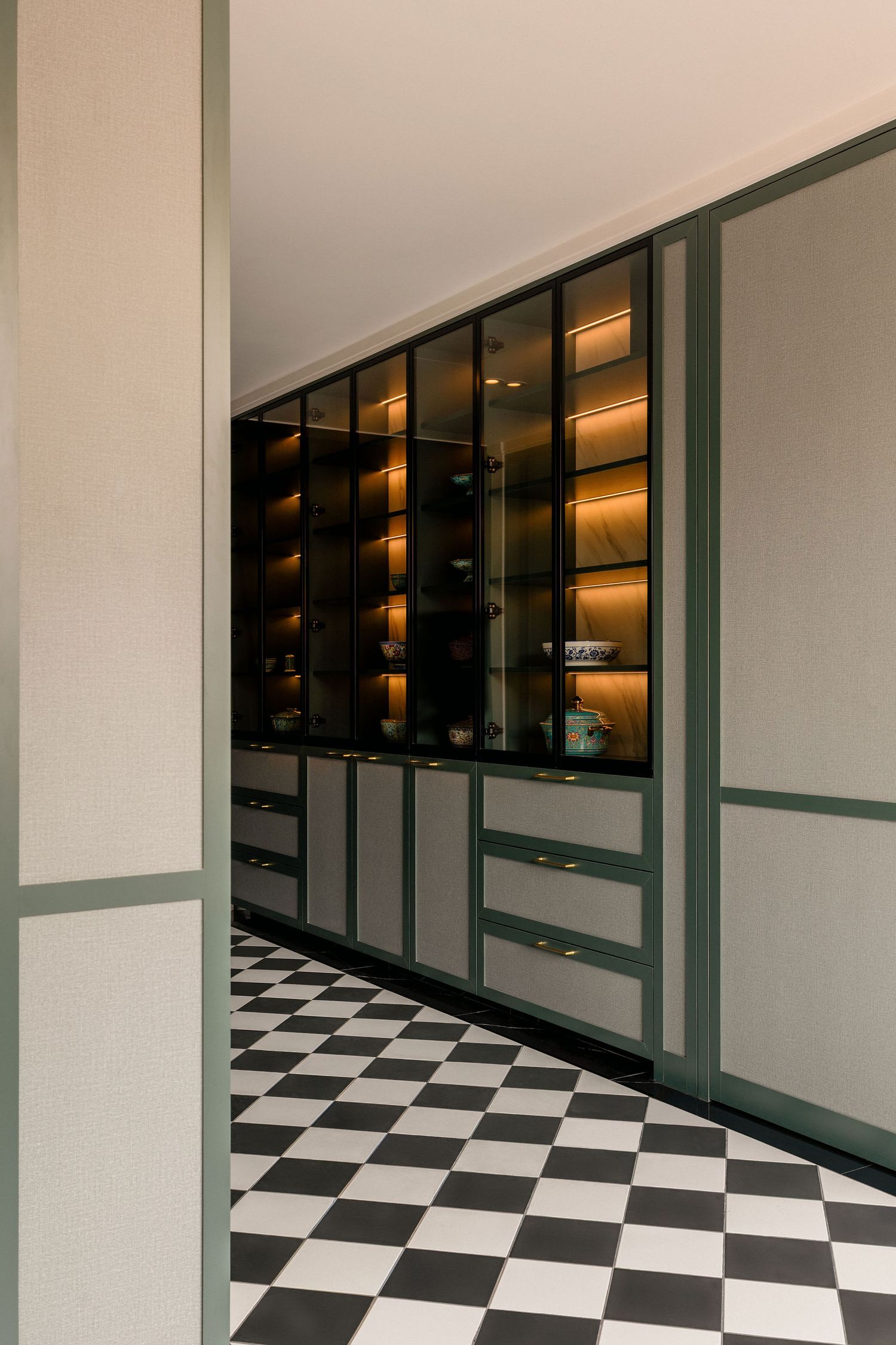 Built-in cabinetry with glass-front display shelves, warm lighting and sage green trim above a black and white checkerboard tile floor.