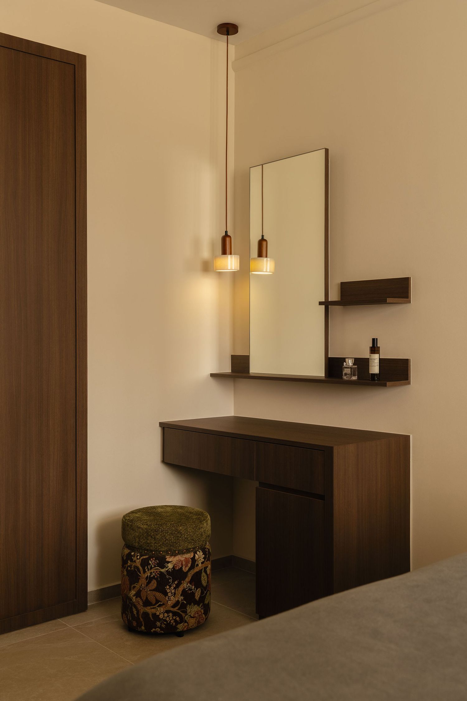 Modern bedroom corner with a walnut wood vanity desk, wall mirror, floating shelf, pendant lights and a floral velvet pouf stool.