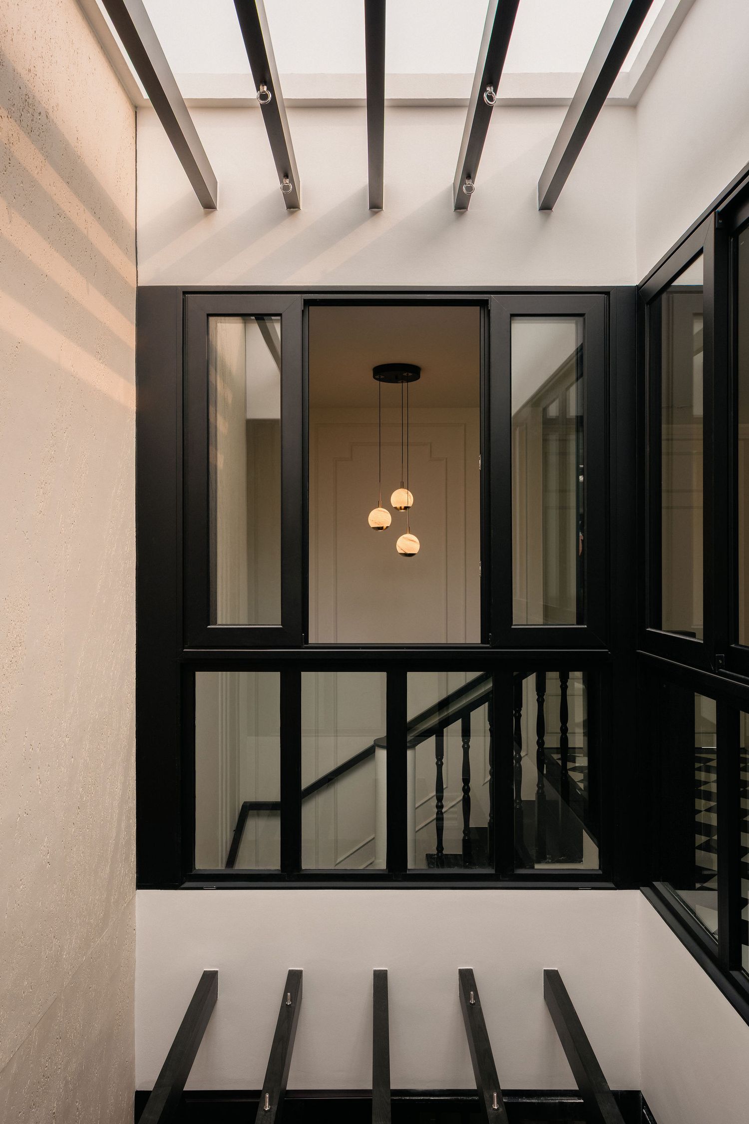 Modern stairwell with black-framed glass windows, a pendant light cluster, and sunlit white walls under an open slatted skylight.