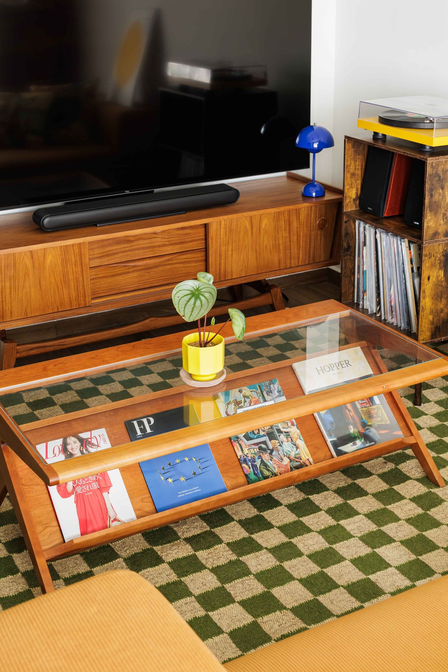 A wooden display shelf holding magazines and books sits on a green checkered carpet beneath a desk lamp.