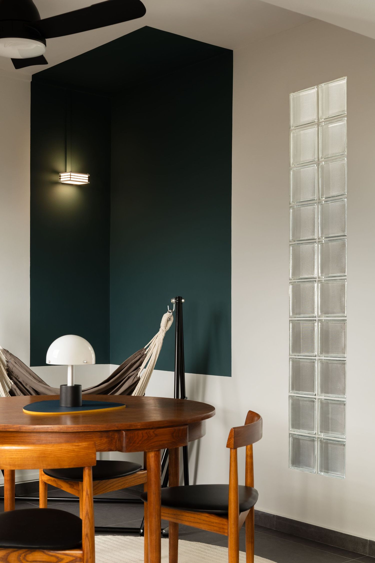 Modern dining area with dark green accent wall, wooden round table, and glass block windows providing natural light.