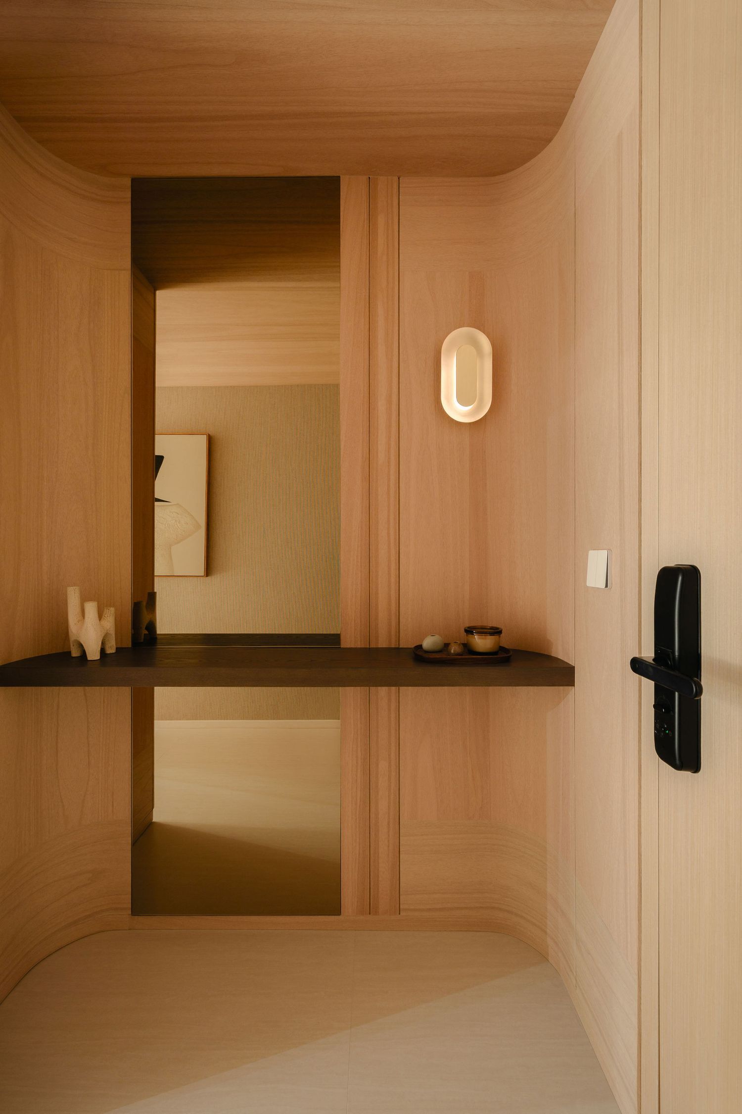 Minimalist wood-paneled entryway with curved walls, a floating dark shelf, mirror, oval wall sconce and modern black smart door lock.
