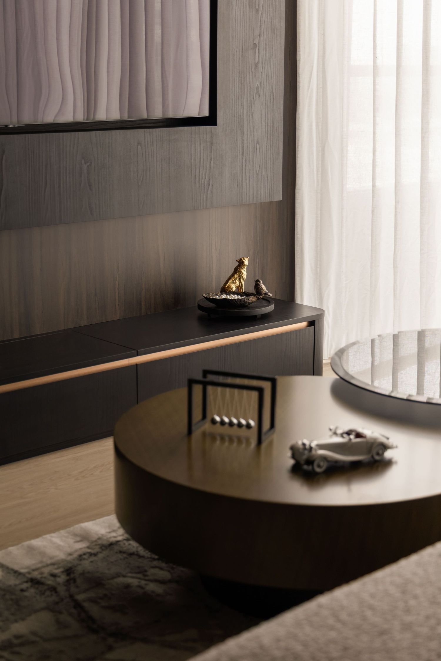 Modern living room with round wooden coffee table, Newton's cradle, and sleek dark TV console against wood-paneled wall.