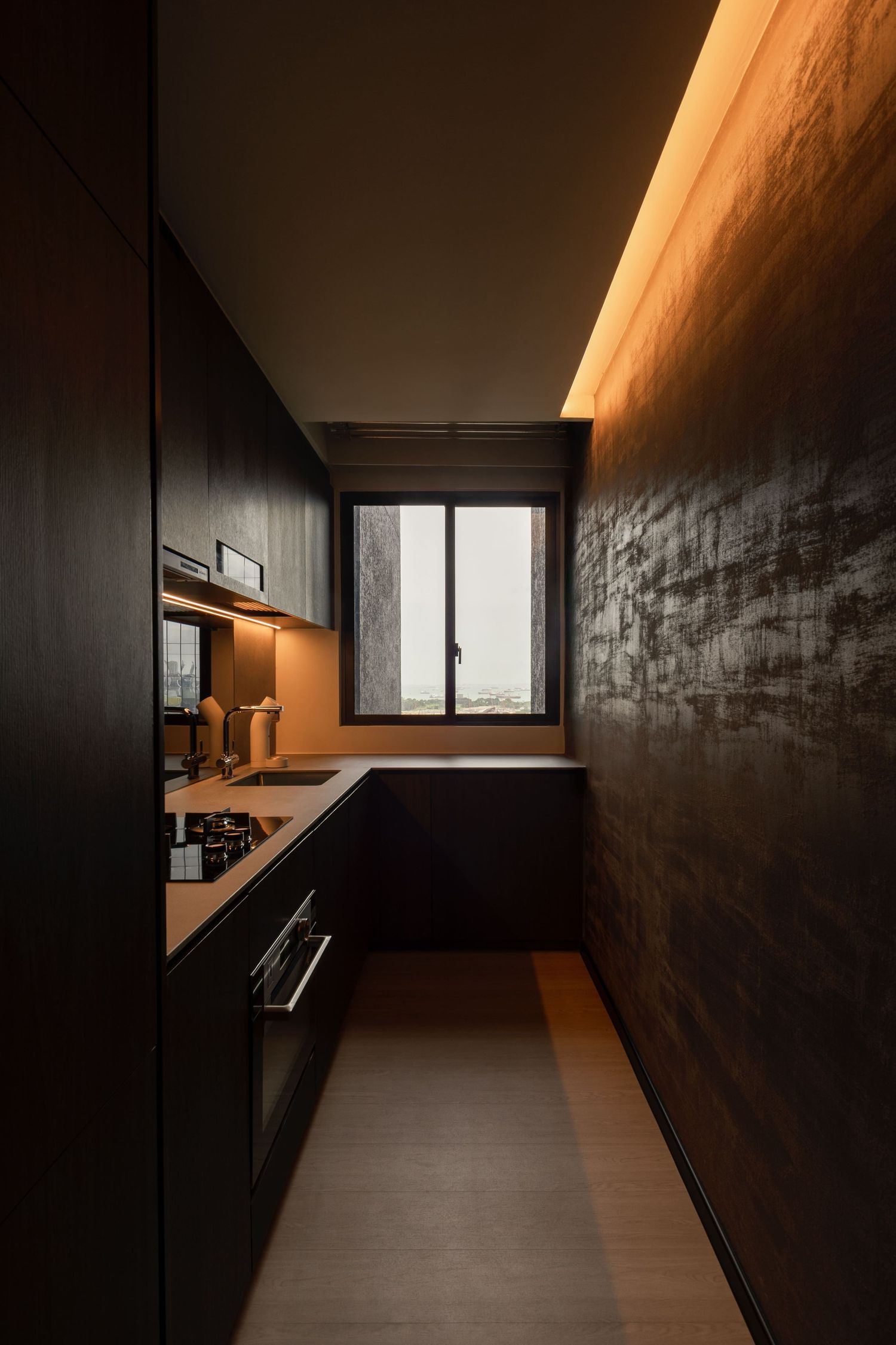 Narrow modern kitchen with dark cabinetry, textured walls, warm cove lighting, and a small window offering city views.