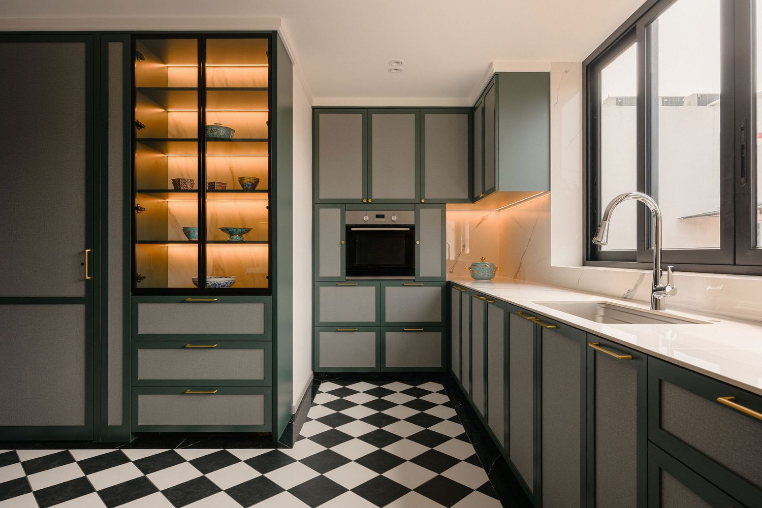 Modern kitchen with sage green cabinetry, black and white checkered tile floor, glass display cabinet, built-in oven and brass hardware.