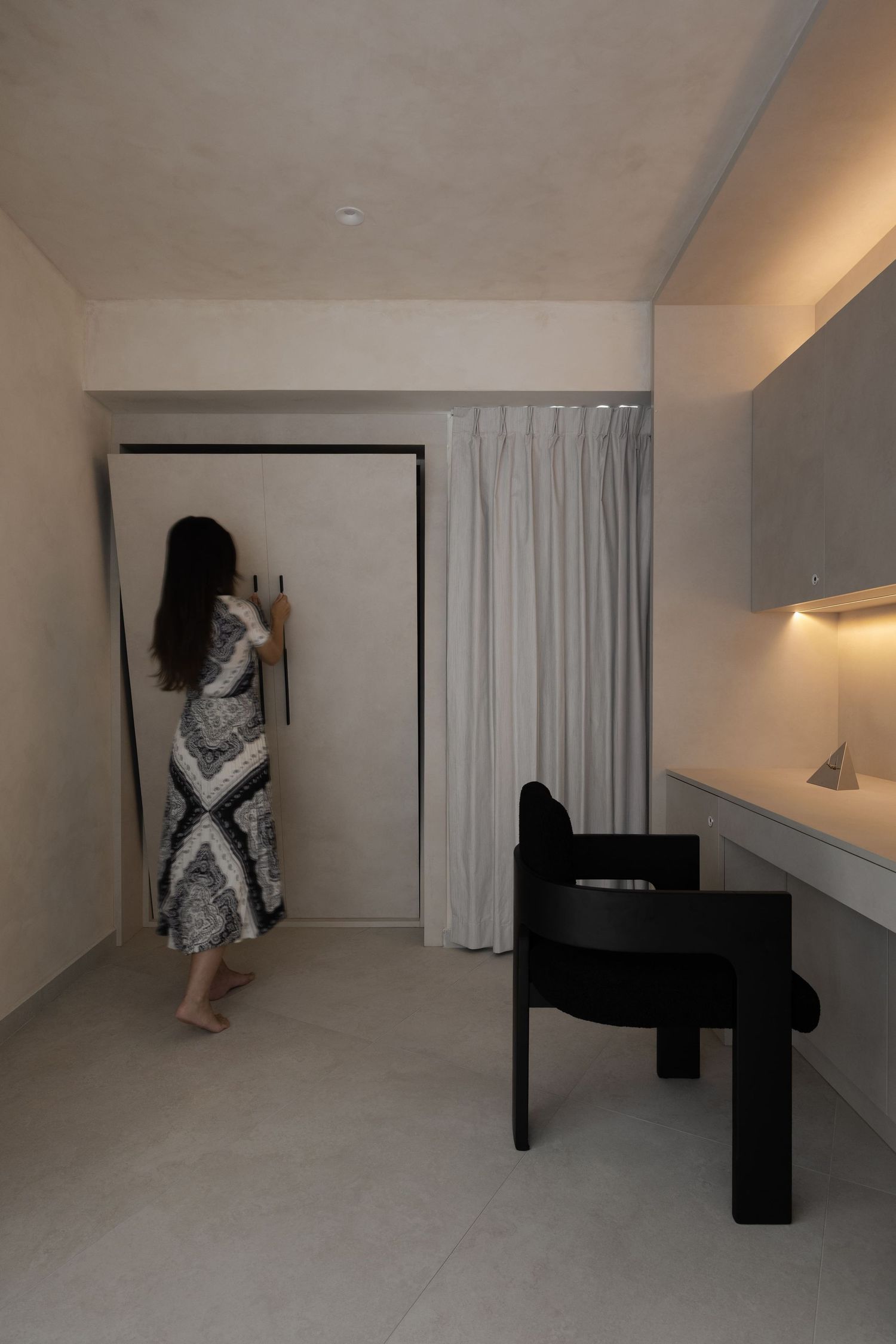 A person in a patterned dress stands near a door in a minimalist room with a black desk and white curtains.