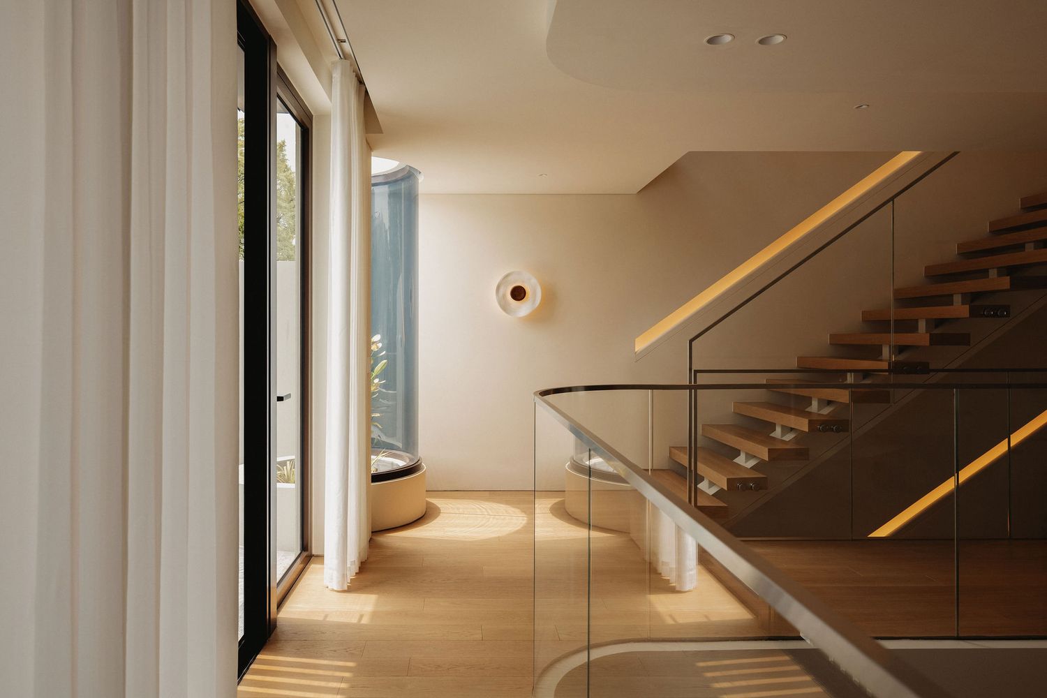 Modern home entryway with glass staircase railing, warm wood floors, and natural light from floor-to-ceiling windows.