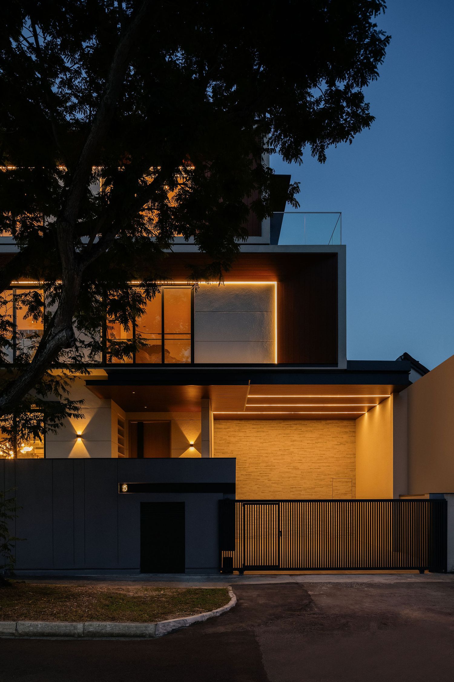 Modern two-story house at dusk with warm LED accent lighting, wood and dark panel cladding, a rooftop terrace, and a slatted wooden gate.