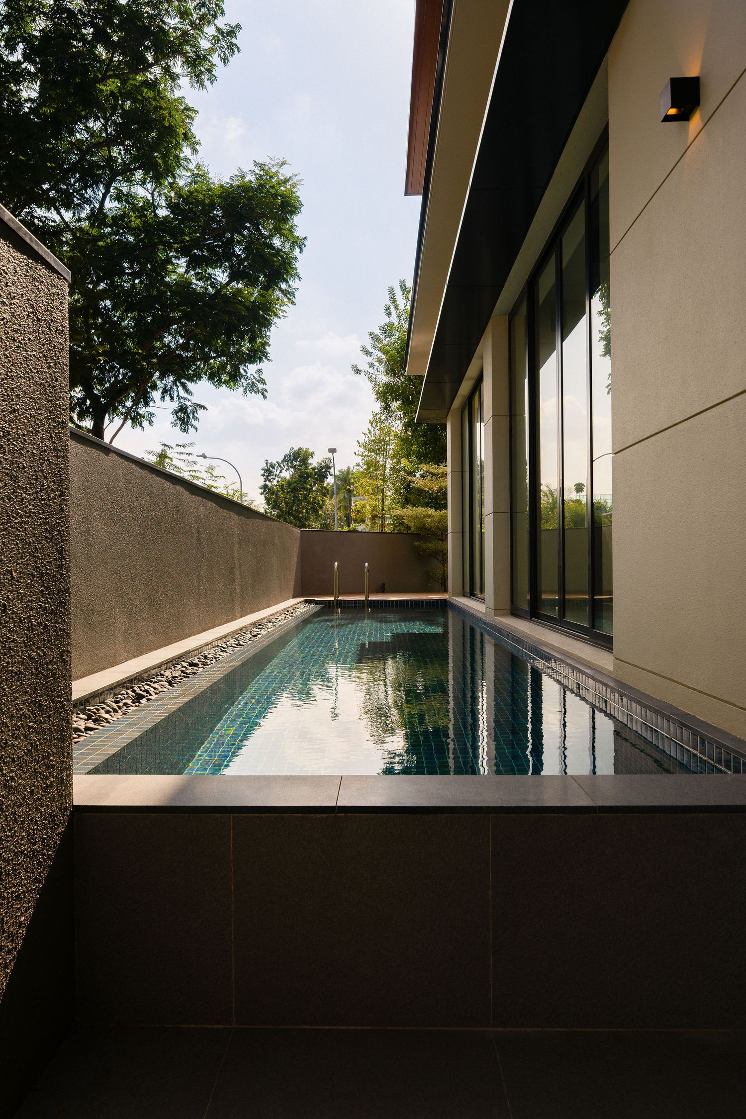 Narrow lap pool with green mosaic tiles beside a modern home's glass facade, textured stone walls and lush trees under a bright sky.