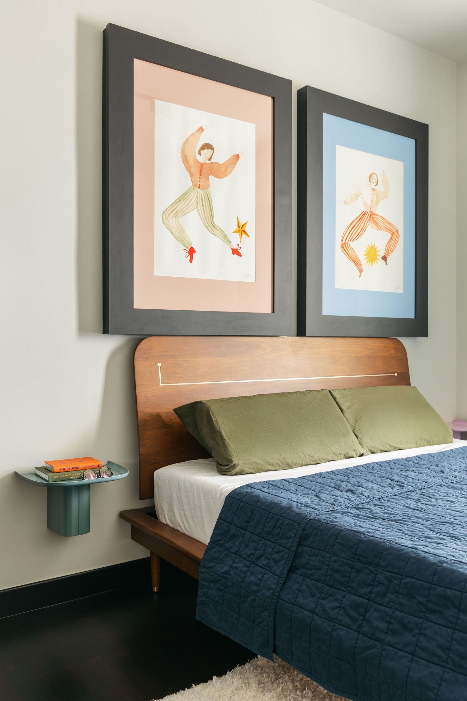 Mid-century modern bedroom with wood headboard, green pillows, navy quilt, and two framed dancing figure artworks above.