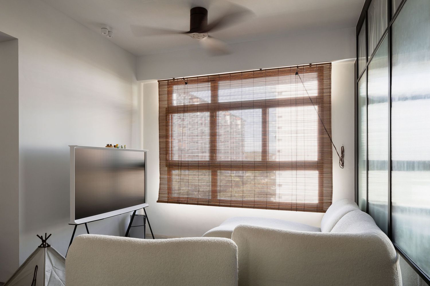 Modern living room with white sofa, mounted TV, bamboo window shade and black ceiling fan in soft natural light.