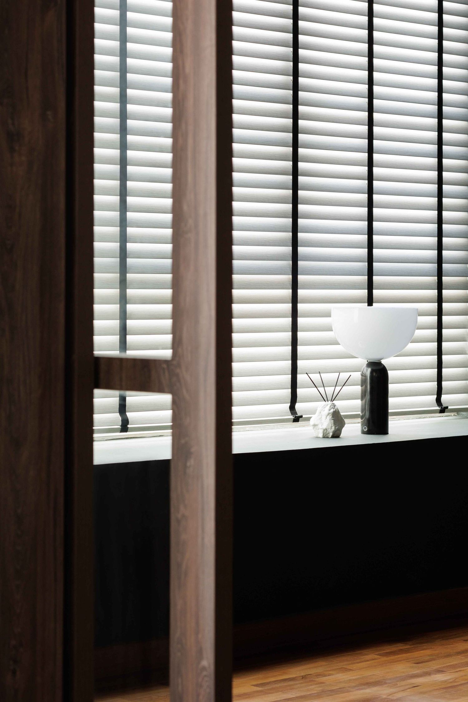 Sunlight streams through white horizontal window blinds casting shadows onto a dark wooden wall surface.