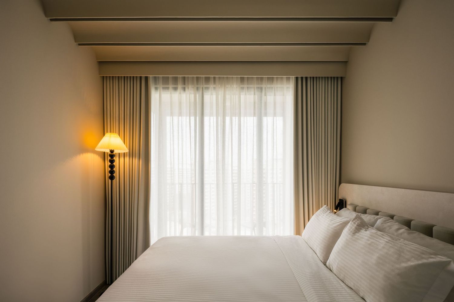 Elegant hotel bedroom with white bedding, sheer curtains, and a warm glowing floor lamp beside a large window.