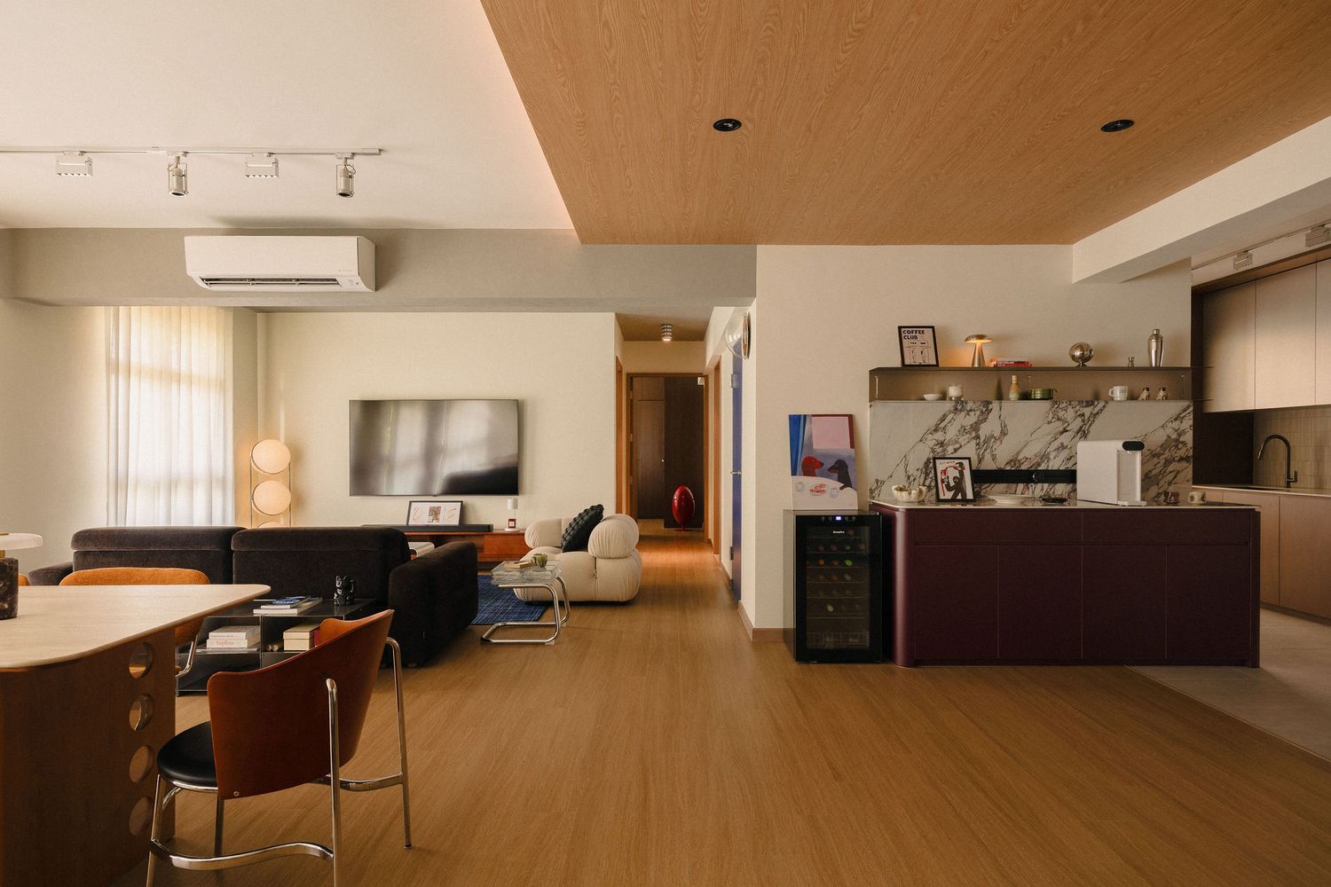 Open-plan apartment interior with wood flooring, a dark navy kitchen island, a living area with sofa and TV, and a warm timber ceiling.