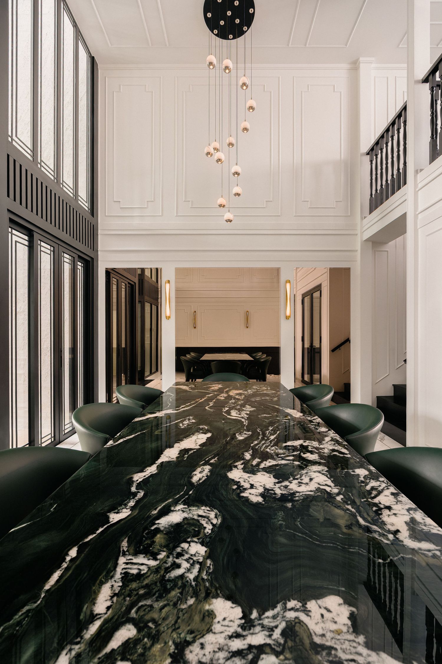 Luxury dining room with a black and white marble table, green velvet chairs, a pendant chandelier and tall paneled walls.