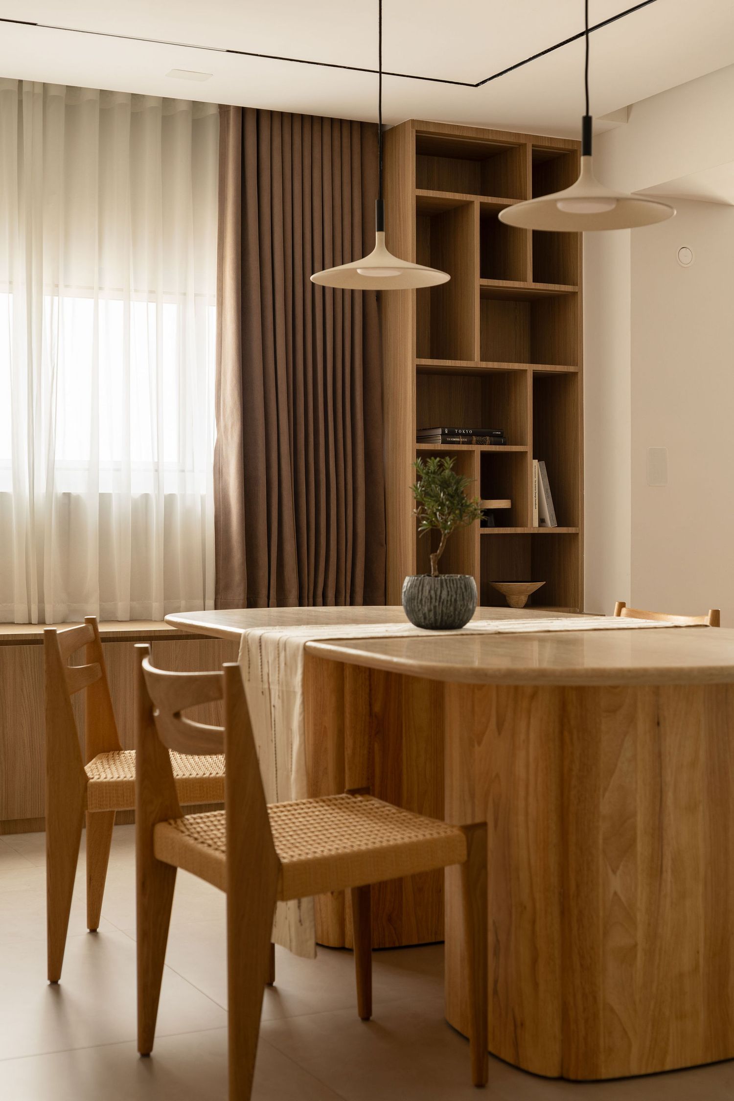 Modern dining area with wooden table, rattan chairs, pendant lights, and built-in shelving unit against brown curtains.