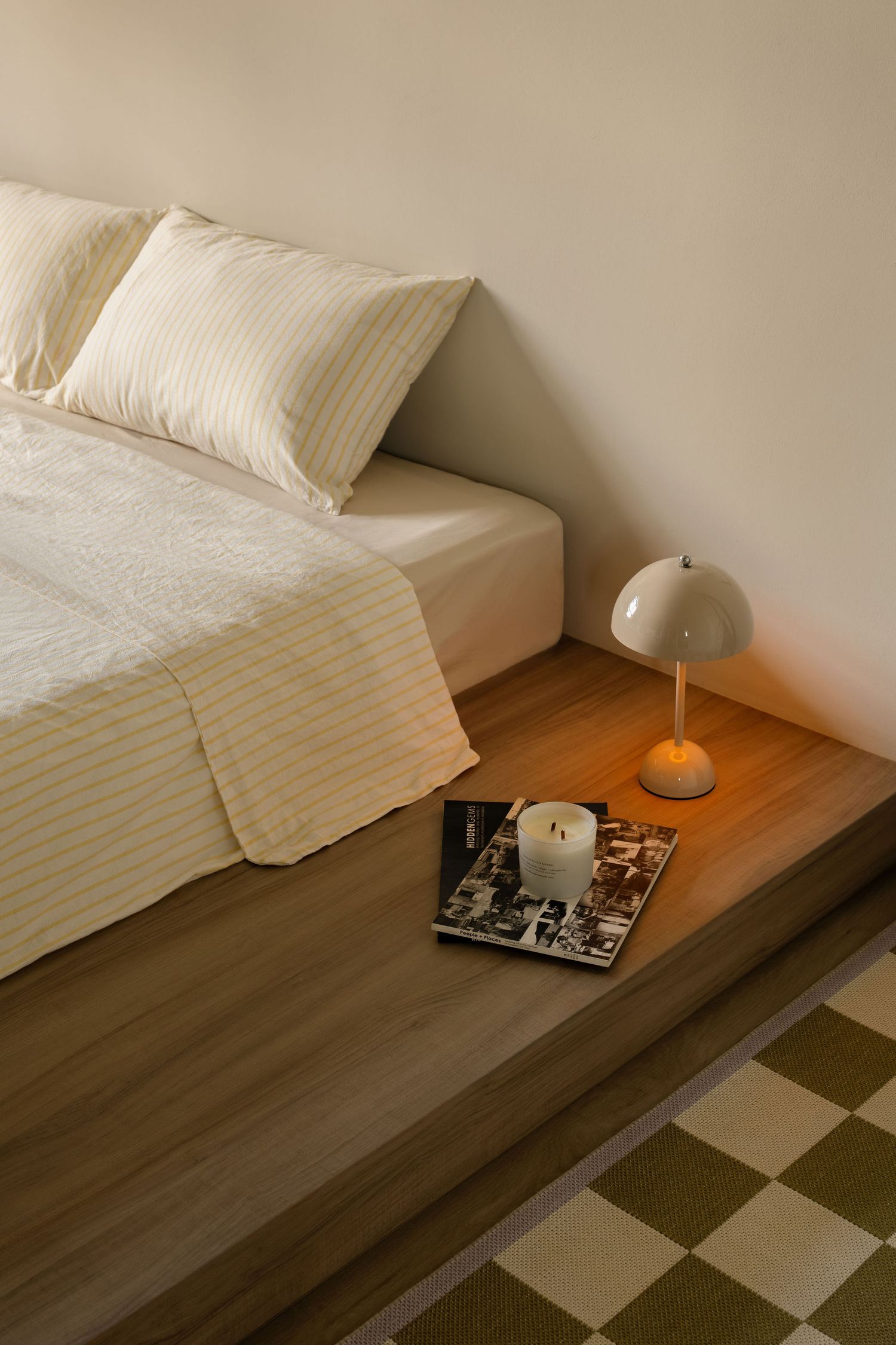Minimalist bedroom with striped bedding, wooden platform bed, glowing mushroom lamp, lit candle, and checkered rug.