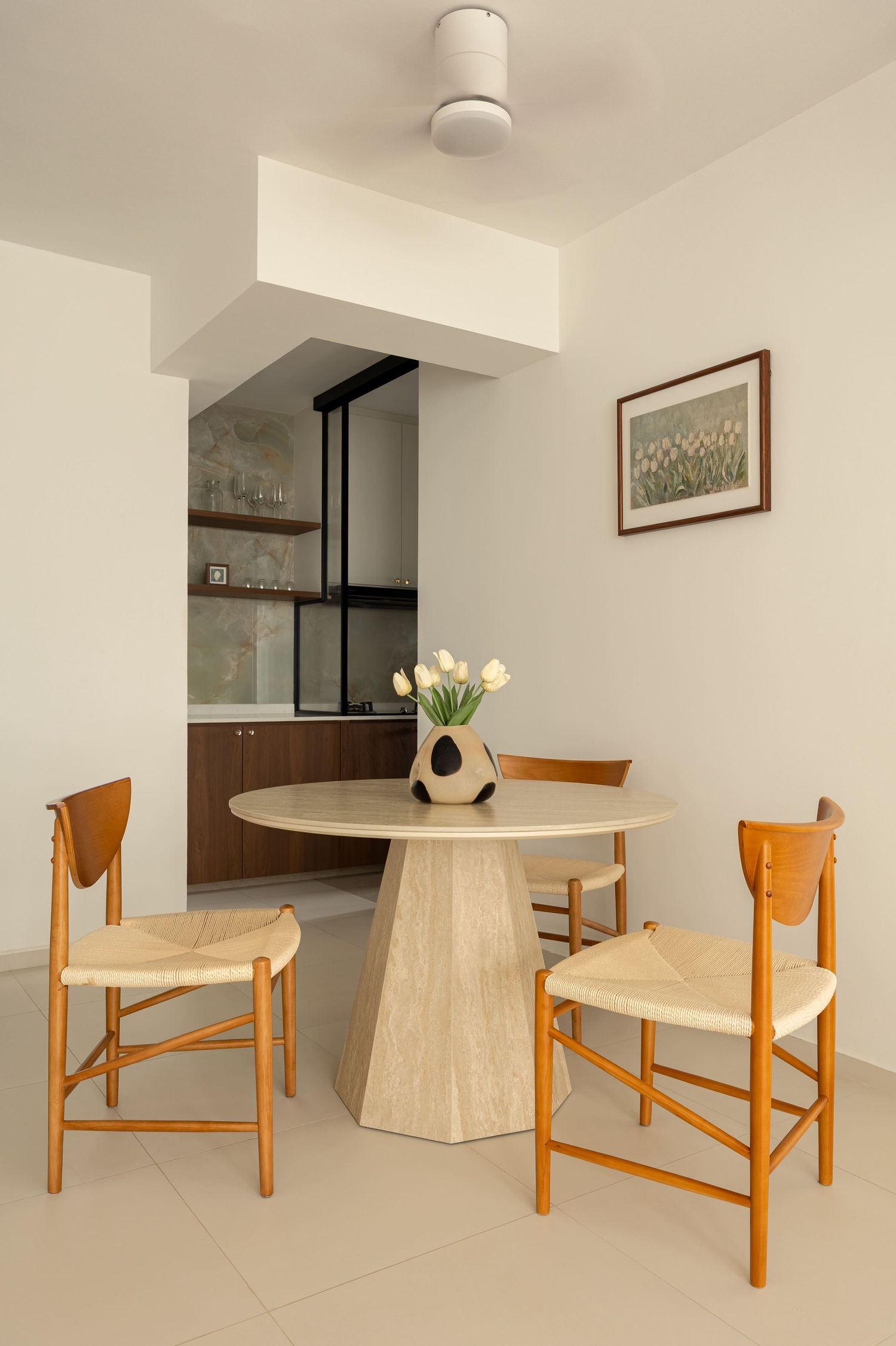 Modern dining area with round travertine table, wooden chairs, tulip vase, and open kitchen backdrop in neutral tones.