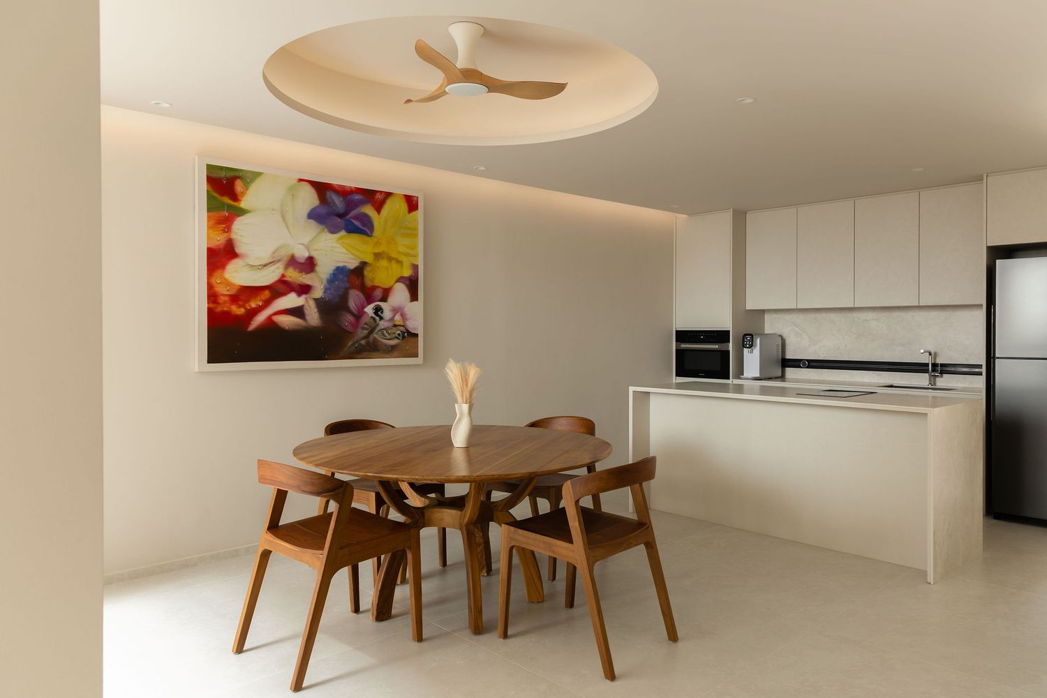Modern dining area with round wooden table, chairs, colorful floral artwork, ceiling fan, and sleek white kitchen in background.