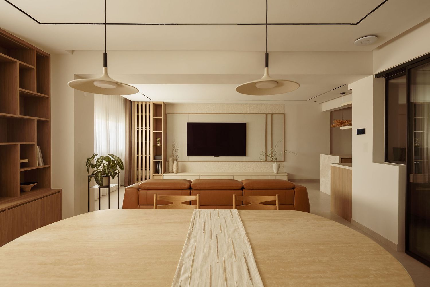 Modern open-plan living and dining room with warm wood tones, pendant lights, built-in shelving, and a wall-mounted TV.