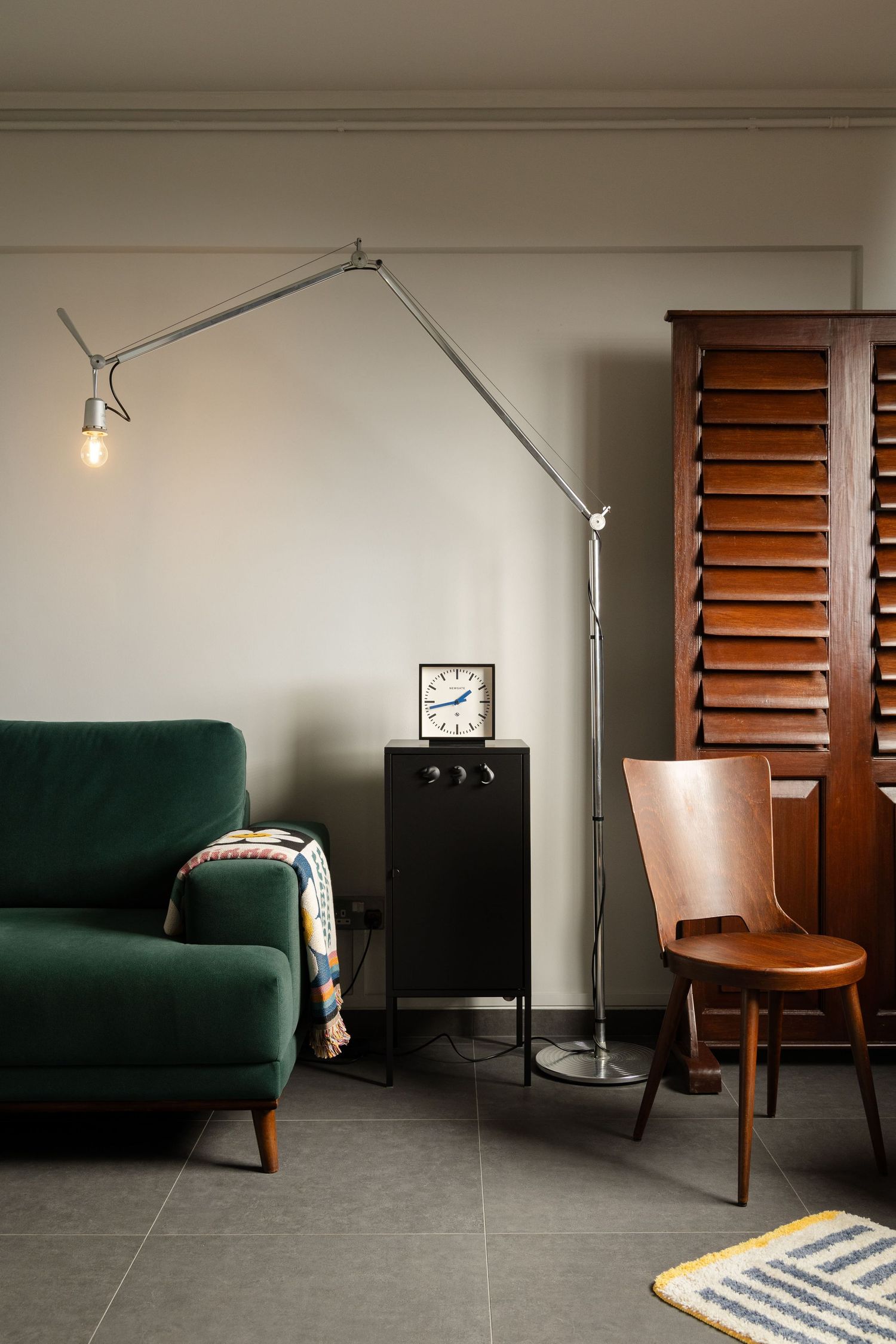 Modern living room corner with emerald green sofa, adjustable floor lamp, wooden chair and slatted window shutter.