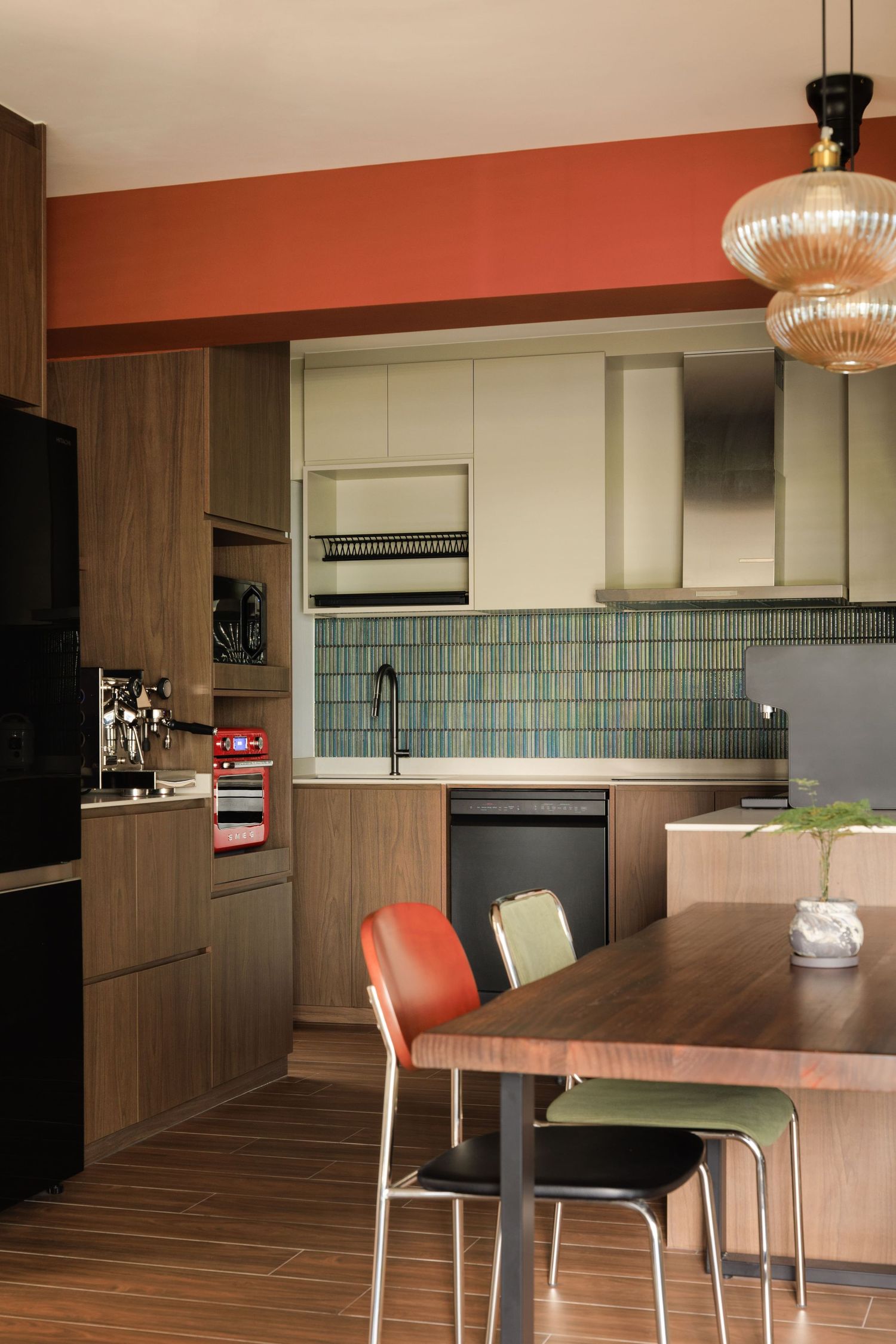 Modern kitchen interior with dark wood cabinets, pendant lighting, and dining area featuring coral-colored accent chairs.