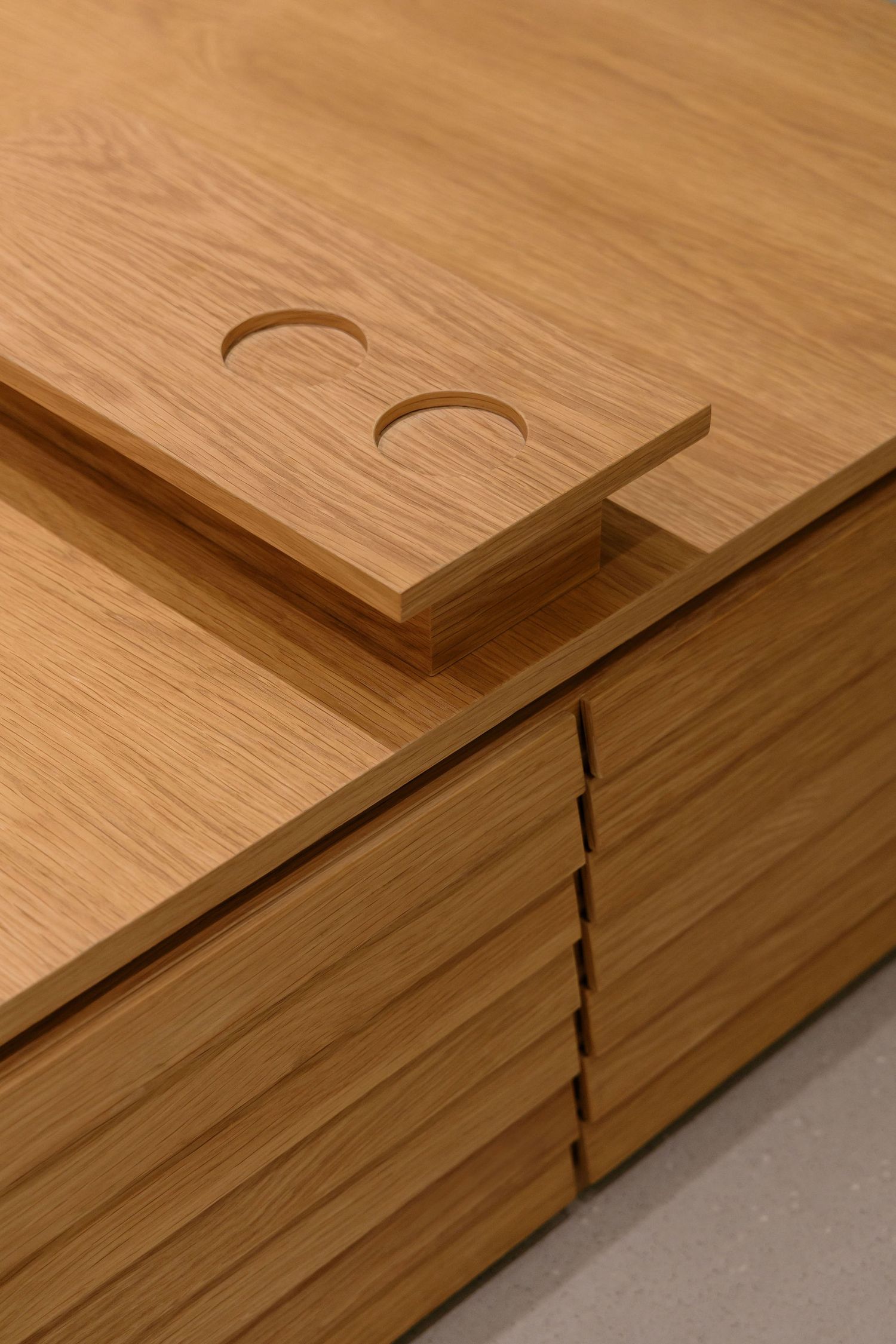 Close-up of a light oak wood cabinet with horizontal slatted drawer fronts and a sliding lid featuring two semicircular finger cutouts.