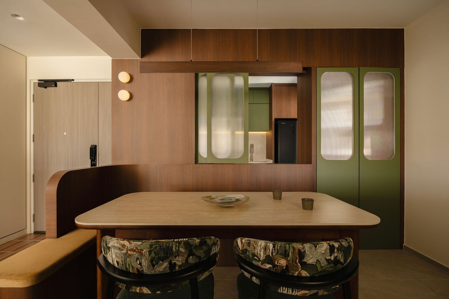 Modern dining area with a wooden table, patterned upholstered chairs, built-in bench, green glass cabinet doors, and warm wood paneling.
