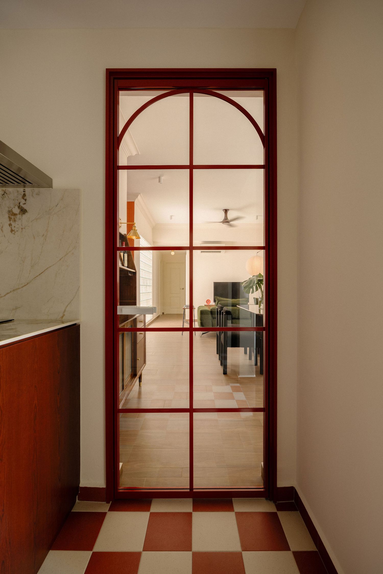 Red-framed arched glass door with grid panes opens from a checkered tile hallway to a bright living area with a ceiling fan.