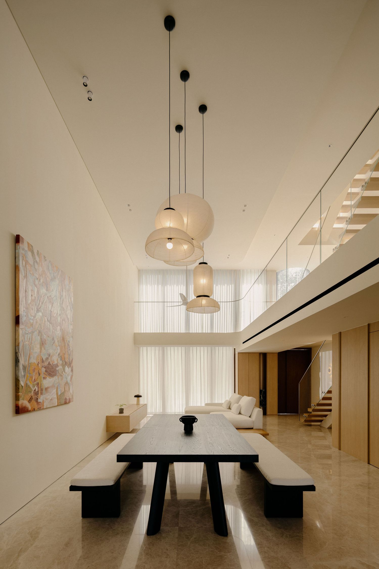 Modern dining room with black table, pendant lights, double-height ceiling, mezzanine level, and sheer curtains in neutral tones.