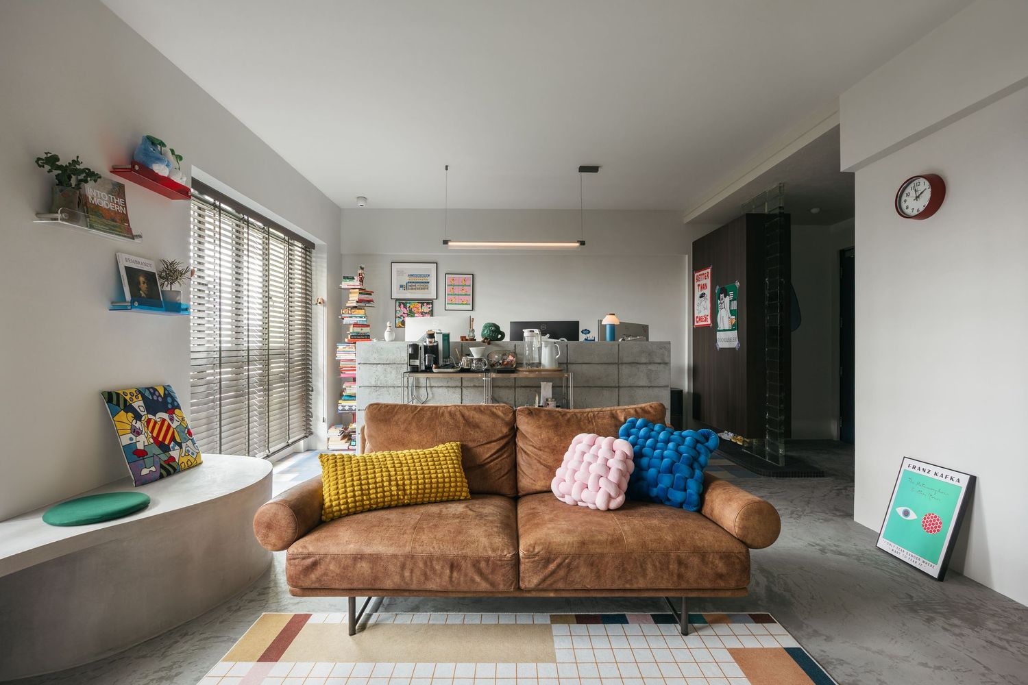Modern living room with brown leather sofa, colorful cushions, striped rug, and open-plan kitchen in neutral tones.