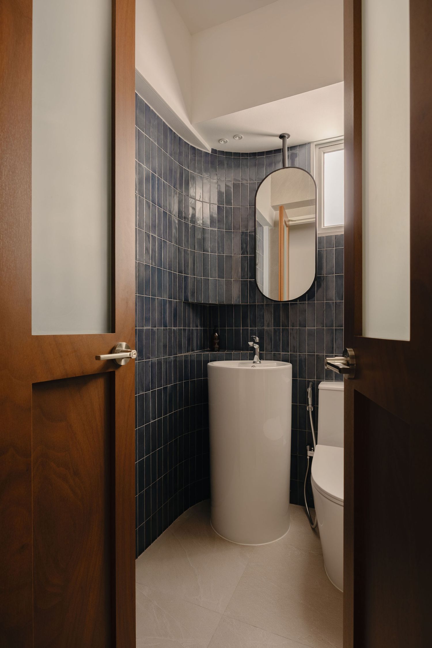 Modern bathroom with blue subway tiles, pedestal sink, oval mirror, and wooden doors.