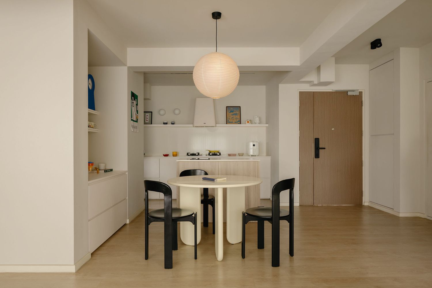 Minimalist dining area with round white table, black chairs, pendant light, built-in shelving, and light wood floors.