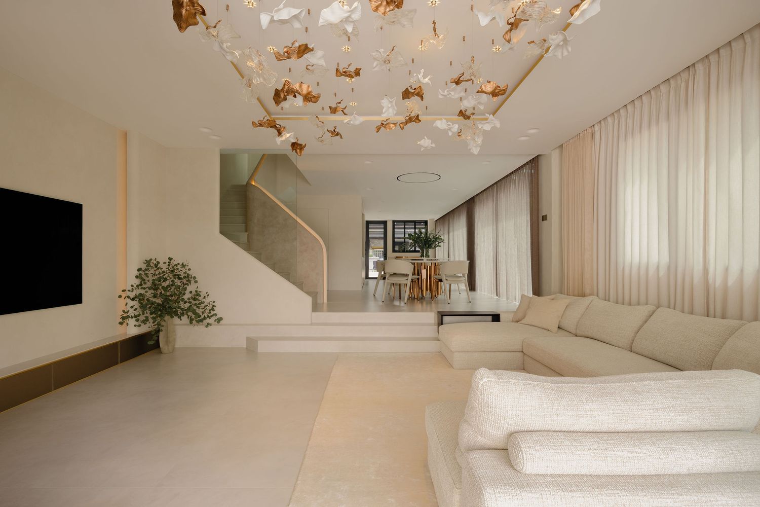Modern white living room with plush sofa, decorative floral ceiling light, staircase, and open-plan dining area beyond.