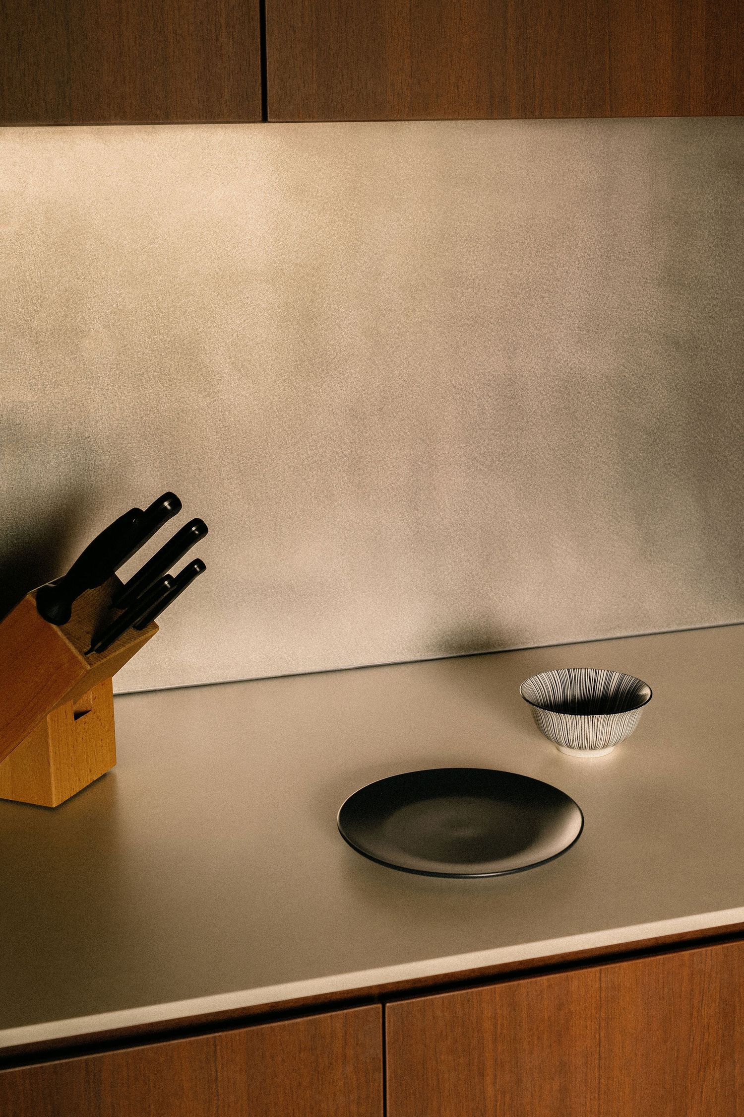 Modern kitchen countertop with a brushed metal backsplash, wooden knife block, black ceramic plate and small ribbed white bowl.