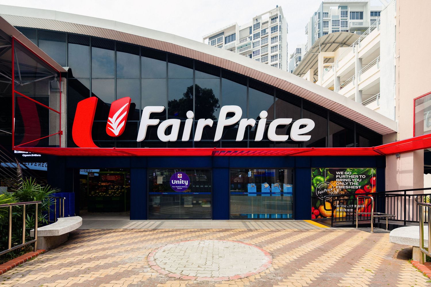 FairPrice supermarket storefront with red and white logo, glass facade, tiled courtyard, bench and fresh produce display near the entrance.