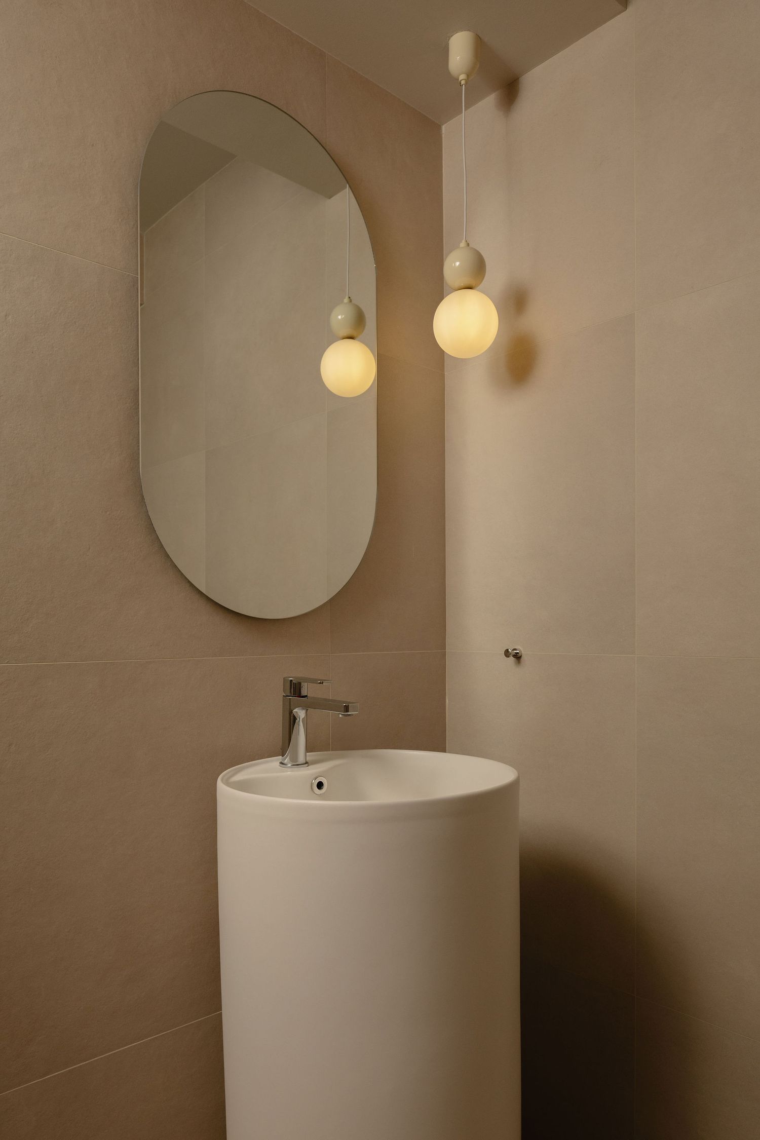 Modern minimalist bathroom with oval mirror, white pedestal sink, and glowing globe pendant lights on beige tiled walls.