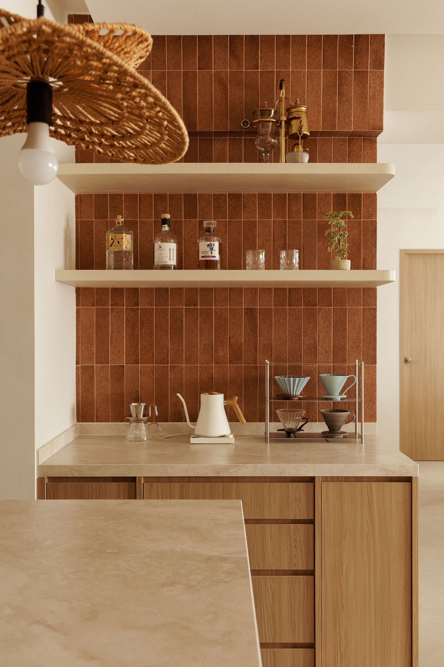 Modern kitchen with terracotta tile backsplash, floating wood shelves, beige countertops, and a woven rattan pendant light.