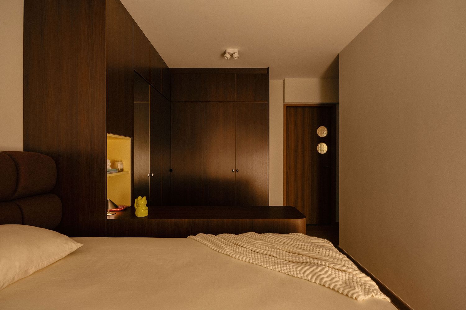 Modern bedroom with warm dark wood paneling, built-in wardrobe, yellow display niche, and a neatly made bed with a textured throw blanket.