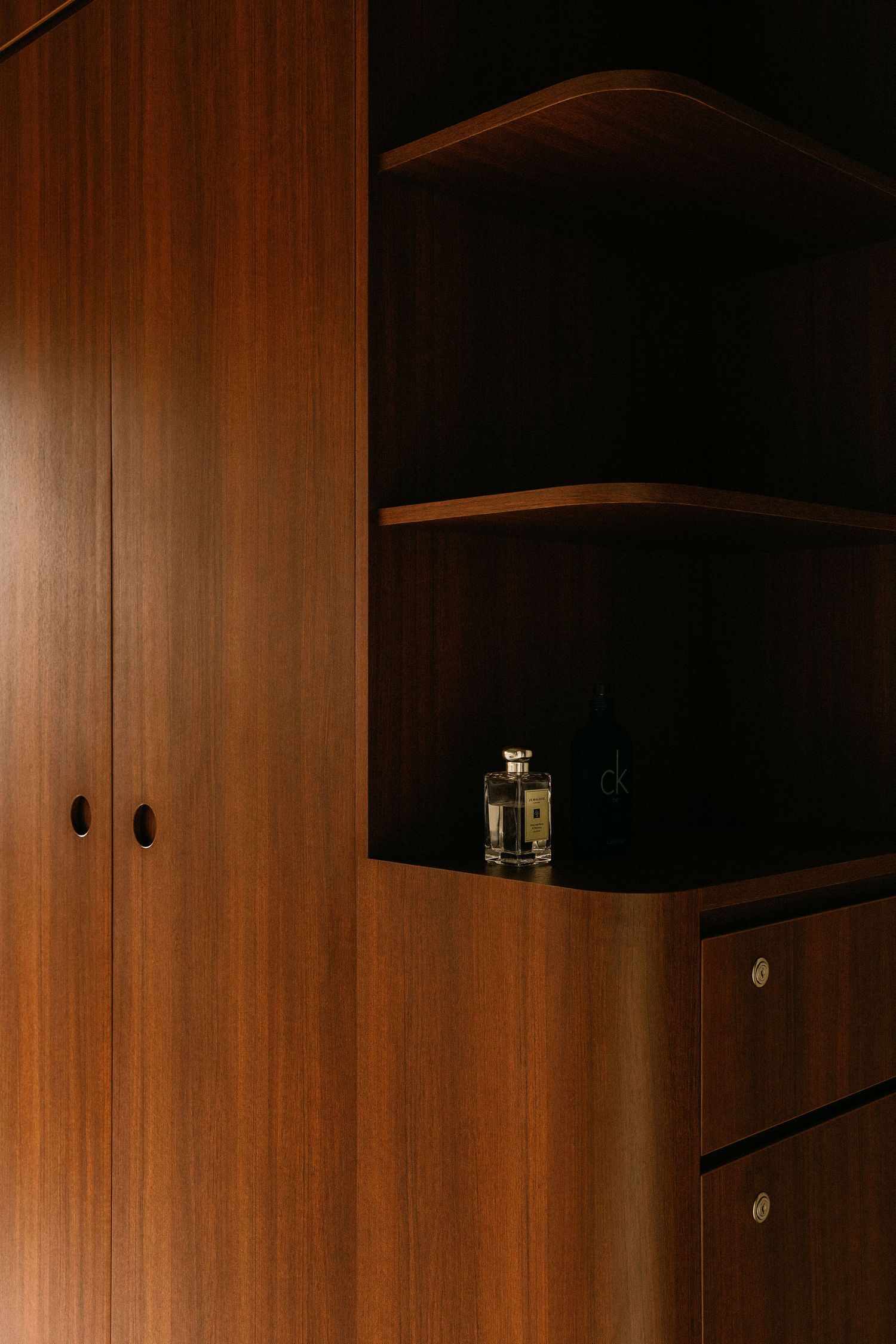 Warm walnut wood cabinetry with curved open shelves, flush doors and brass pulls, displaying a small glass perfume bottle in soft light.