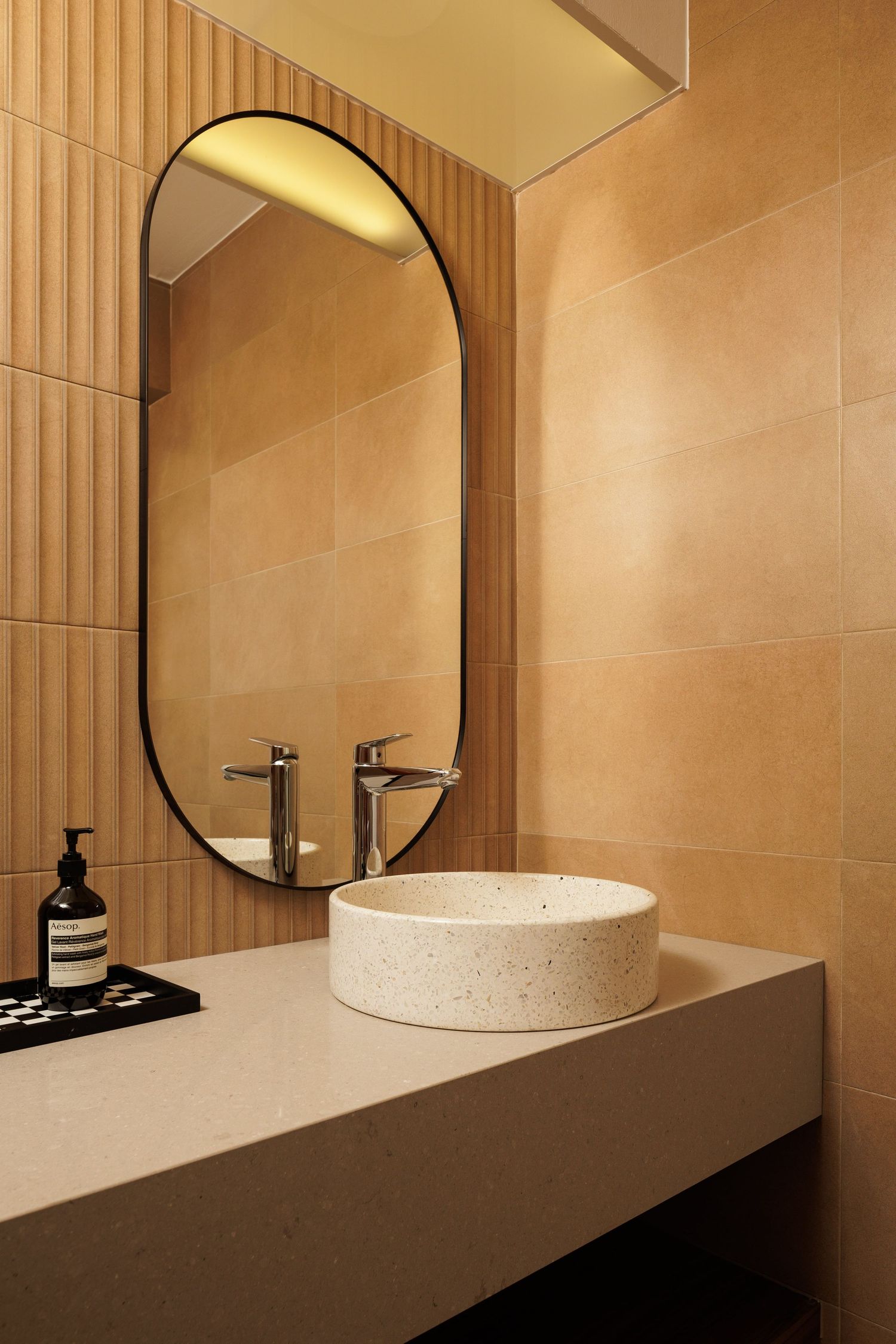Modern bathroom interior with oval mirror, terrazzo vessel sink, and warm beige tiled walls and wooden paneling.