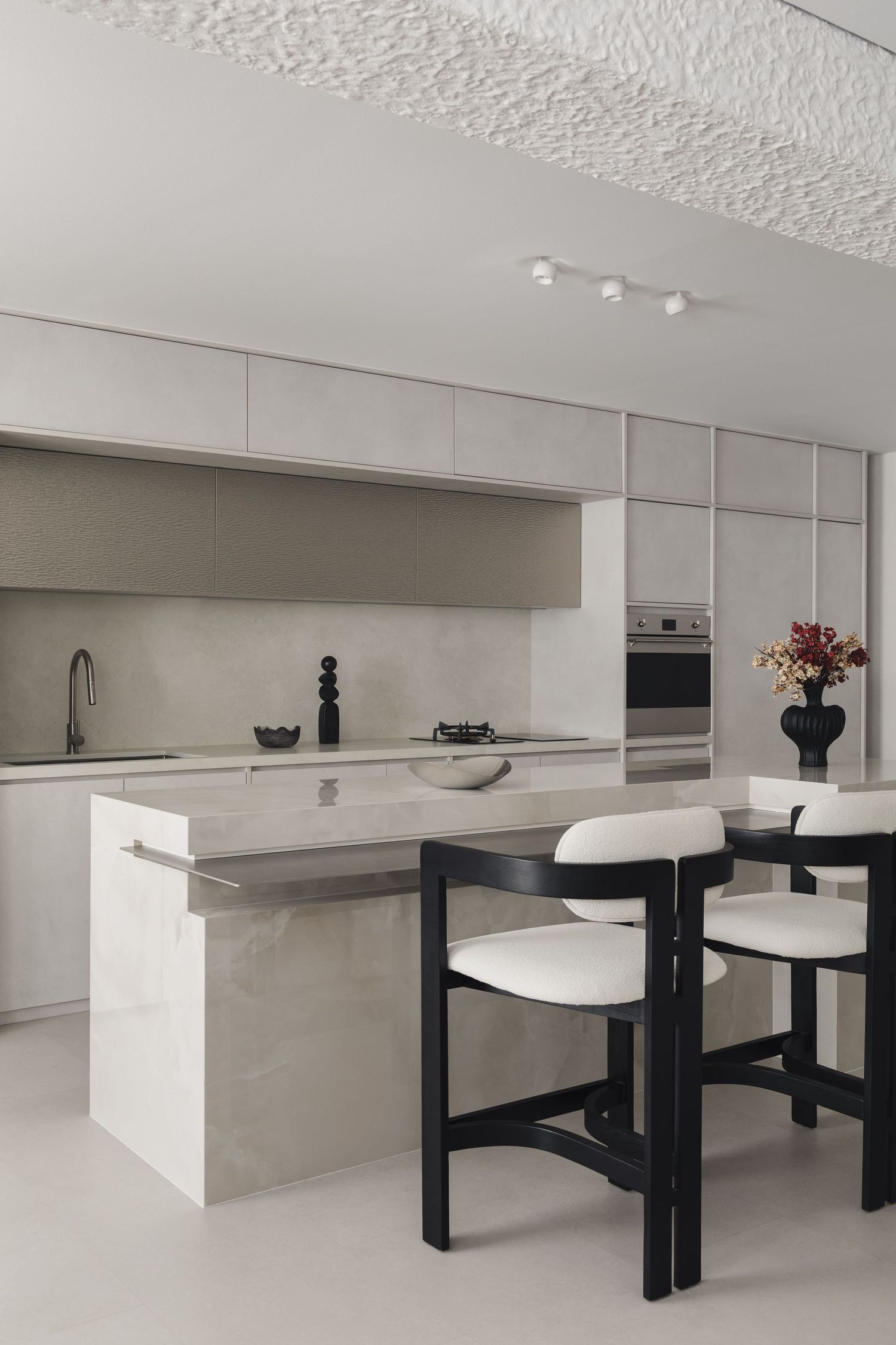 Modern minimalist kitchen with marble island, black bar stools, gray cabinetry, and stainless steel appliances in neutral tones.