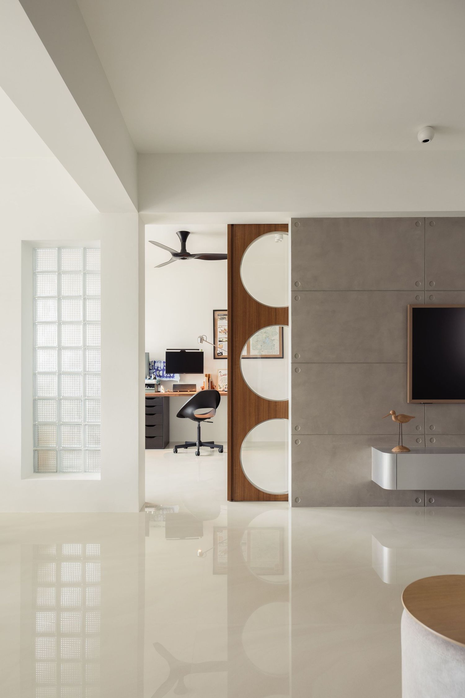 Modern minimalist living space with glossy white floors, concrete accent wall, TV unit, and wooden sliding door with circular cutouts.