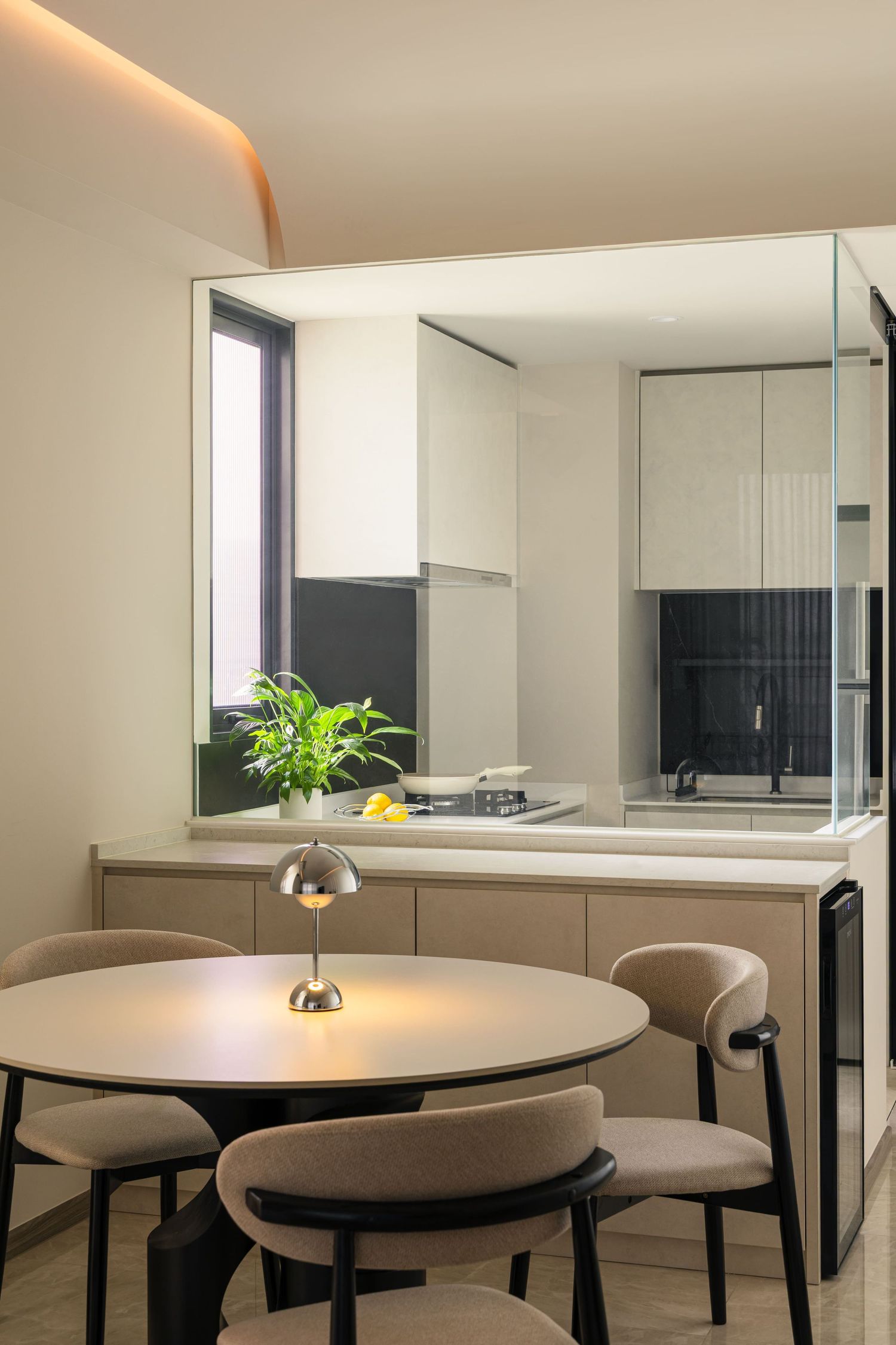 Modern dining area with round table, beige chairs, and small lamp beside open-plan kitchen with glass partition.