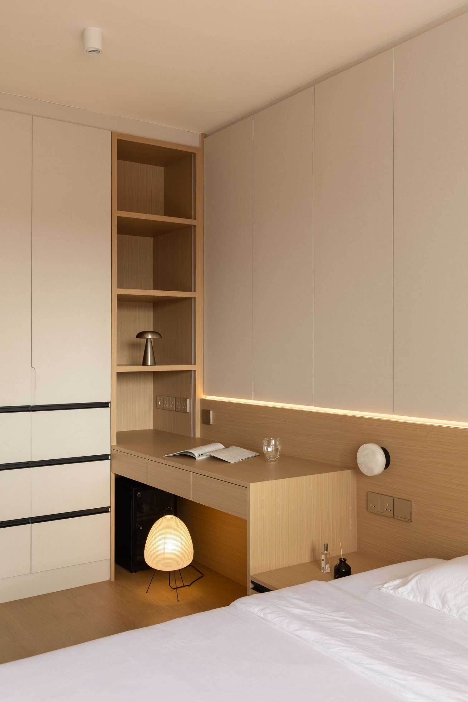 Modern minimalist bedroom interior with built-in shelving, warm lighting, and white bedding against beige walls.