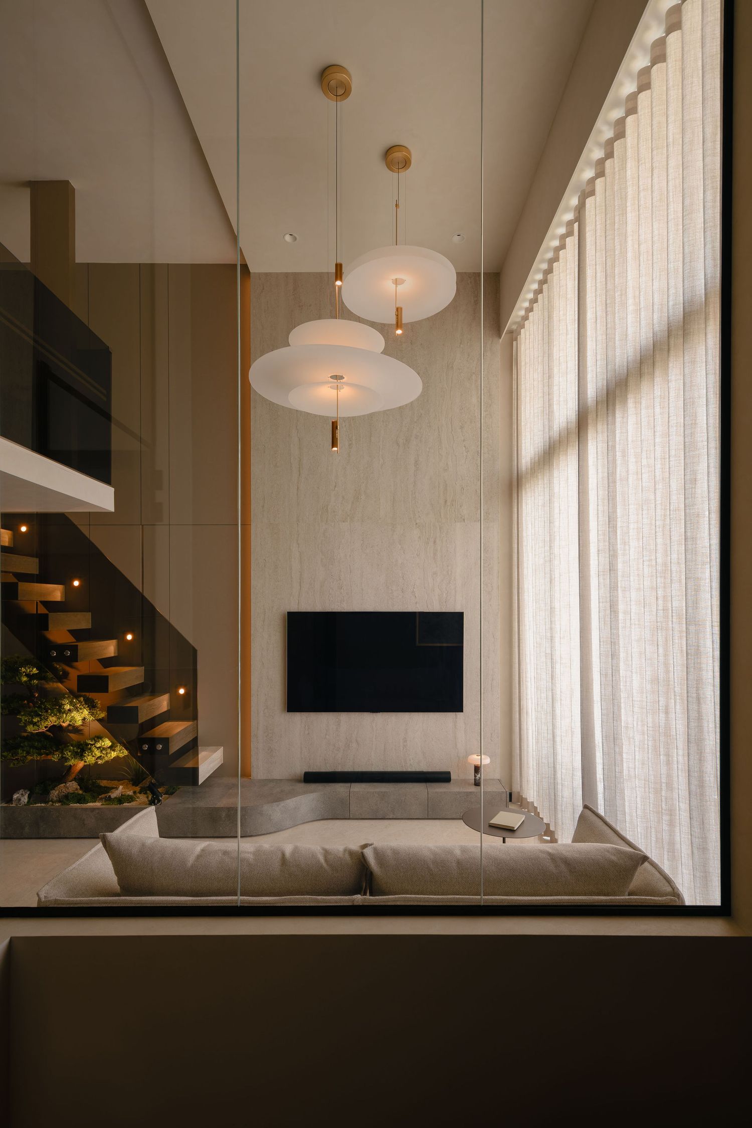 Modern double-height living room with a gray sofa, wall-mounted TV, pendant lights, sheer curtains and a floating wooden staircase.