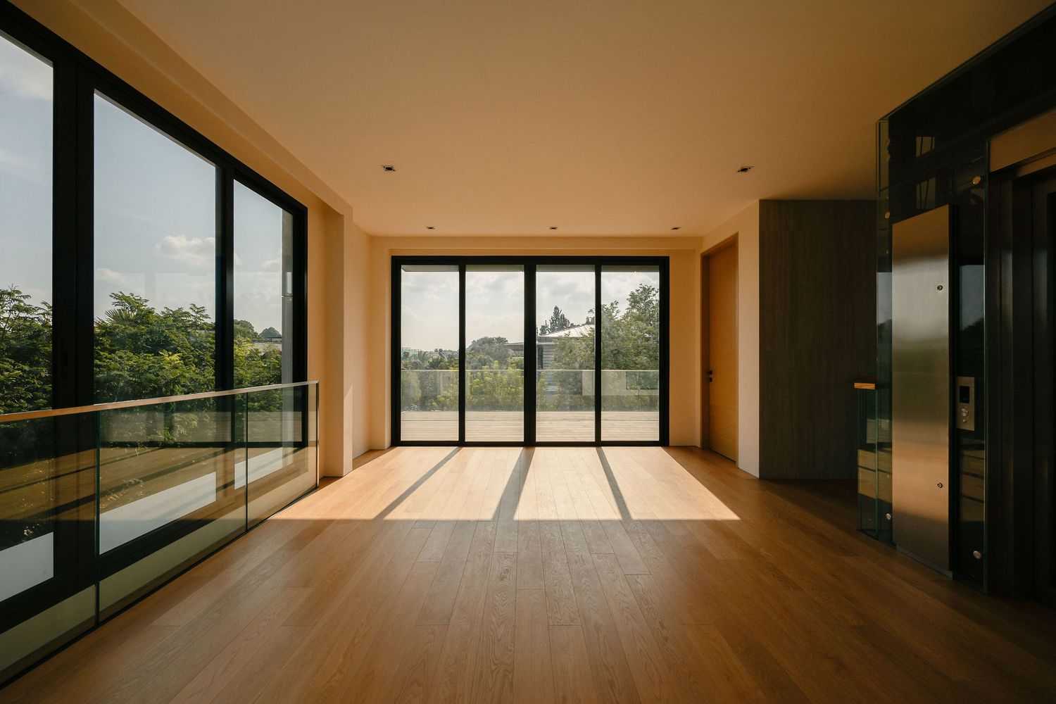 Spacious modern living room with hardwood flooring, floor-to-ceiling glass windows, glass balcony railing, and an elevator, overlooking lush