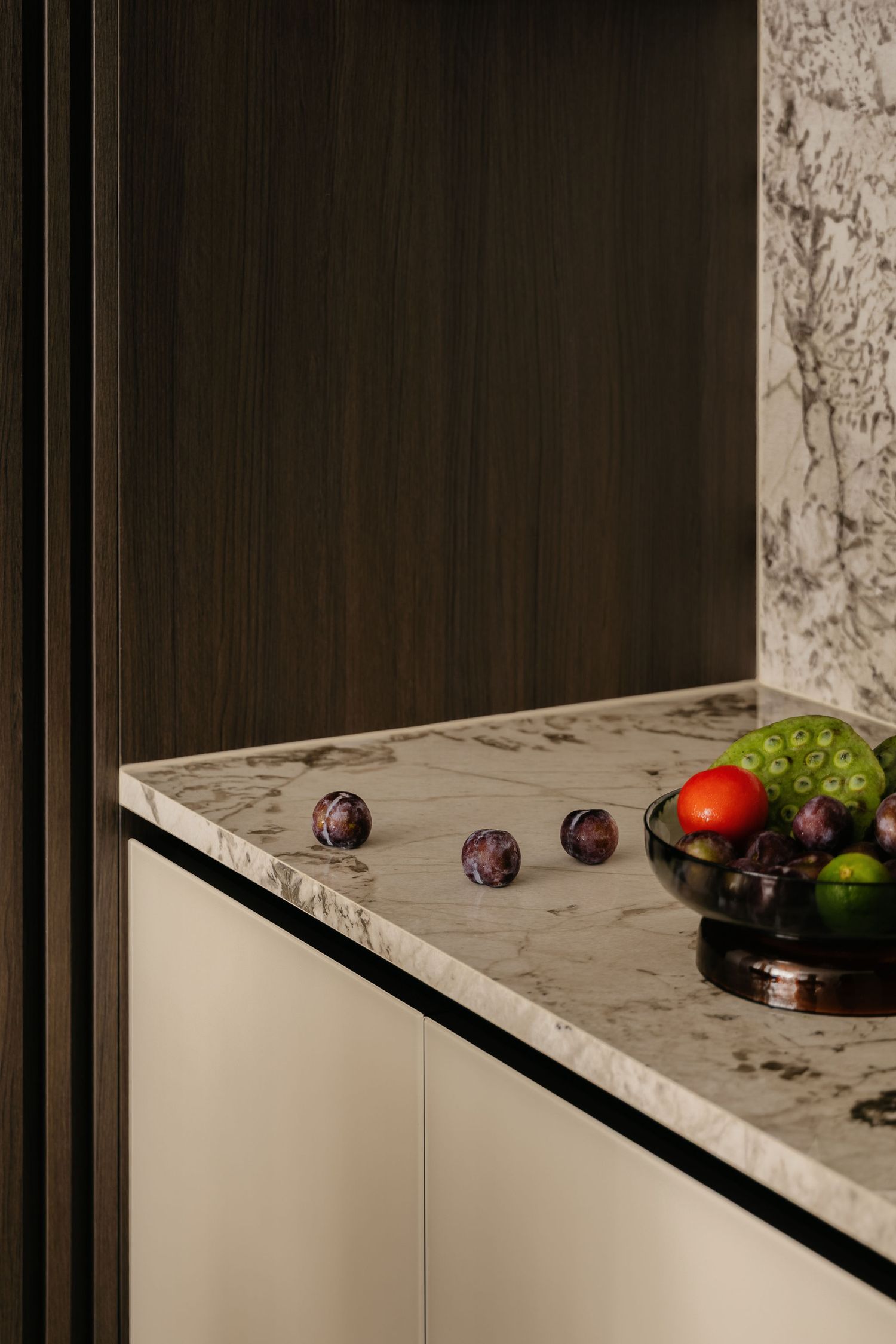 Marble kitchen countertop corner with scattered plums and a dark bowl of fruit, beside cream cabinets and dark wood paneling.