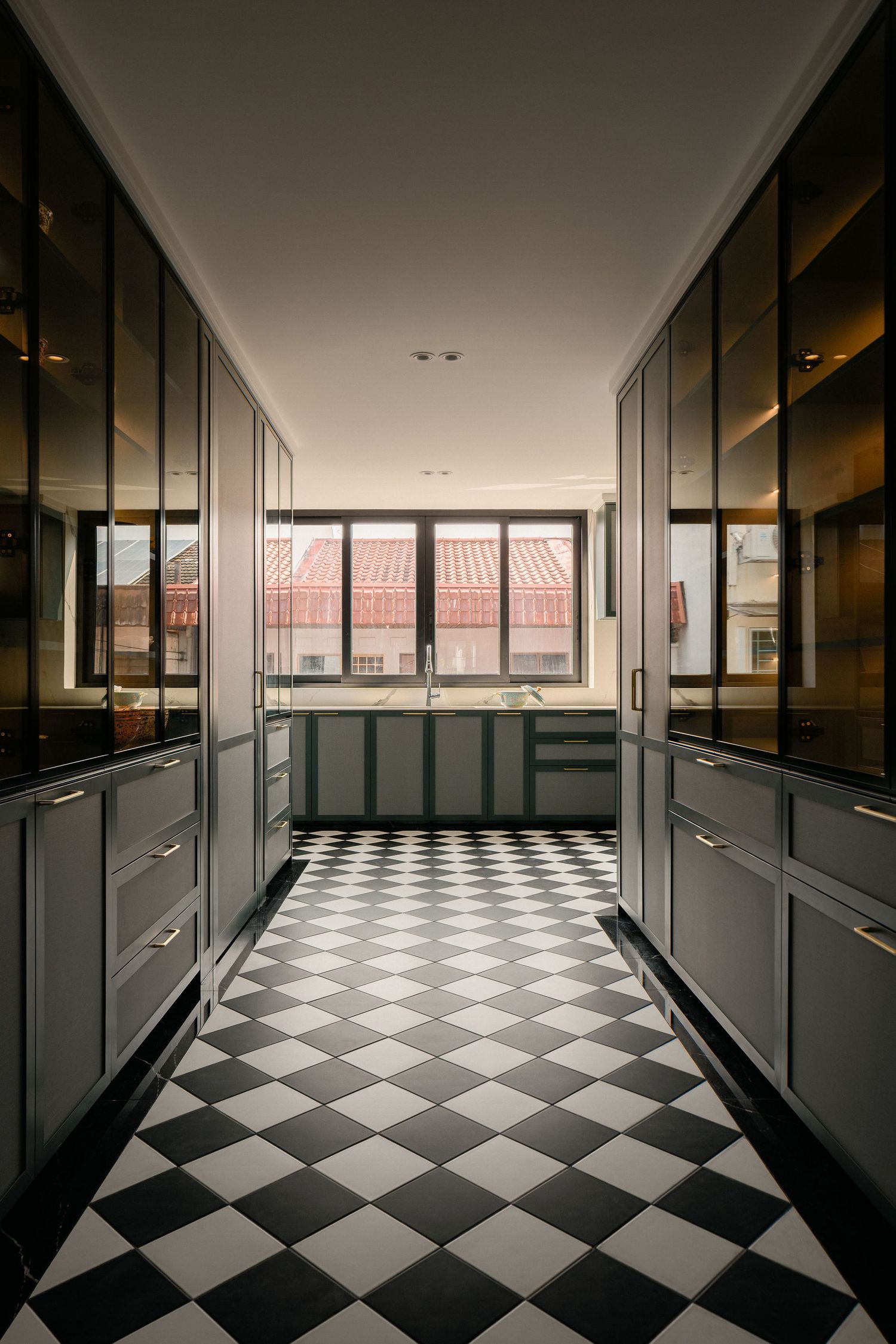 Modern galley kitchen with gray cabinetry, glass-front upper cabinets and a black and white checkered tile floor leading to a window.